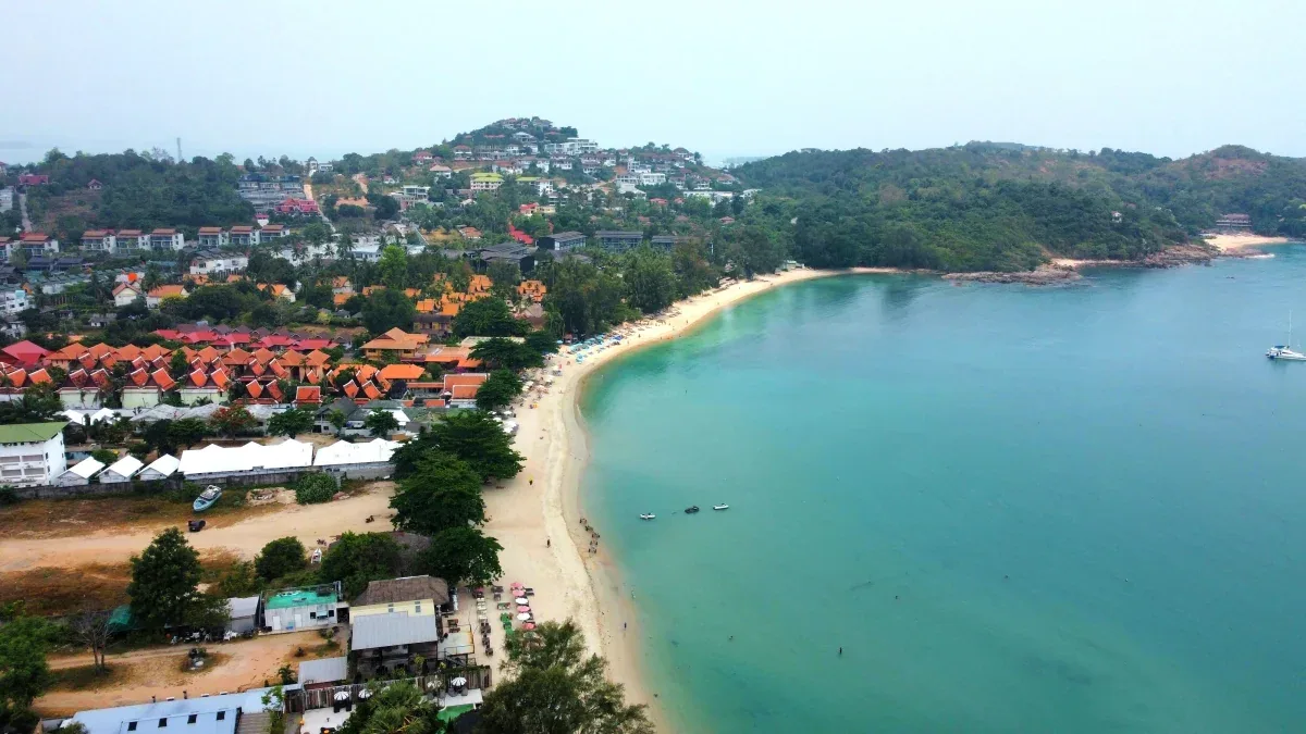 Aerial view of a pristine curved bay in Koh Samui with turquoise waters, golden sandy beach, red-tiled resort buildings, and hillside homes surrounded by lush tropical forest